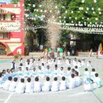 Group of students in white uniforms sit in a large circle on a school courtyard during a festival with confetti falling and festive banners overhead Sarathi school habsiguda hyderabad
