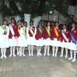 Line of schoolgirls in maroon blazers and white uniforms posing outdoors wearing colorful sashes designating prefects under a large tree in a school courtyard Sarathi school habsiguda hyderabad