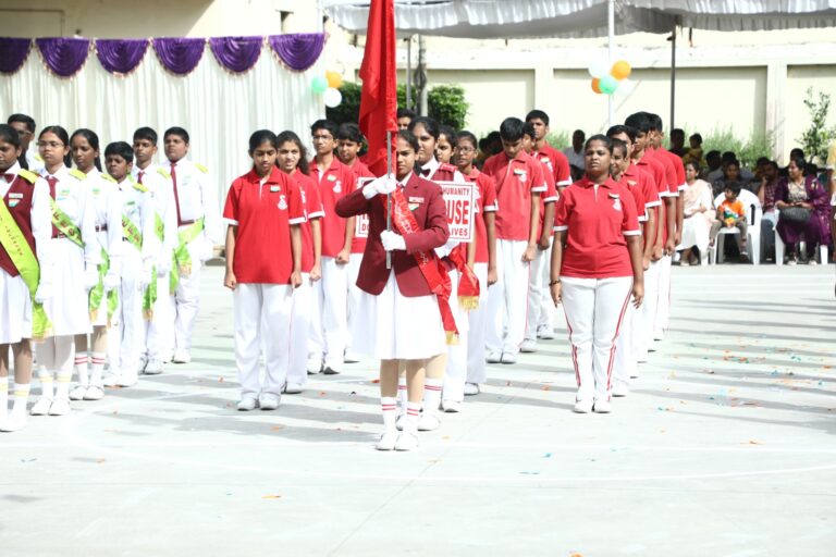 School parade: a girl in a red blazer carries a flag at center; rows of students in red shirts and white pants march, with others in white uniforms on the left; spectators sit in the background.