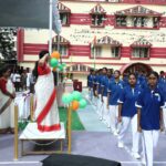 A woman in a white saree with red border speaking at a podium while students in blue uniforms march in formation beside her at a decorated school event Sarathi school habsiguda hyderabad