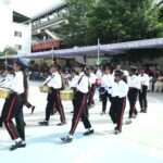 School marching band in white shirts and hats performing a drum procession outdoors with spectators under a canopy tent Sarathi school habsiguda hyderabad
